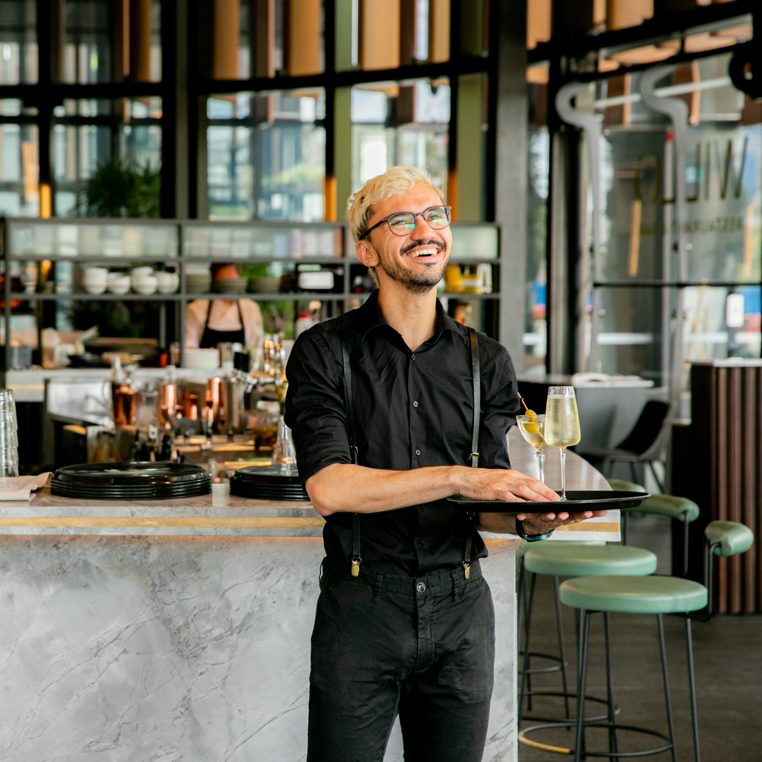 A smiling server with blond hair and glasses holds a tray with a drink. He's in a chic restaurant with a marble bar, set in a modern, stylish ambiance.