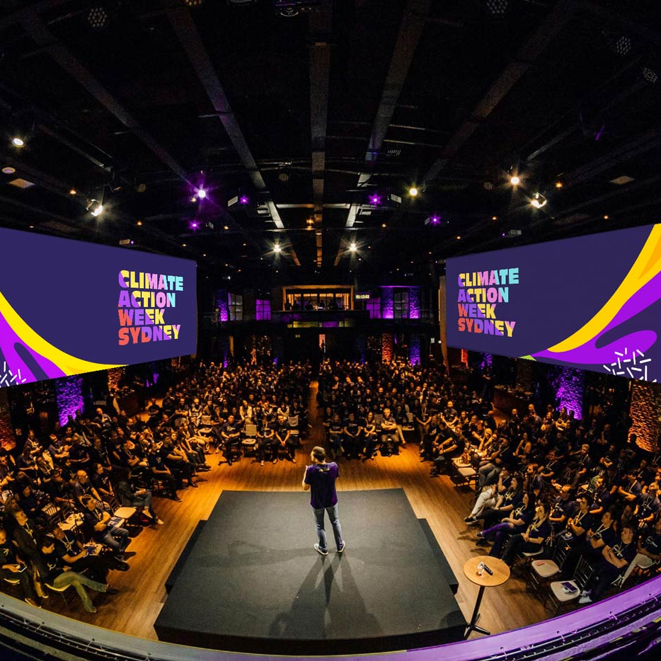 Speaker on stage at Climate Action Week Sydney. Large audience views vibrant graphics on screens, set in a dimly lit auditorium with purple lighting.