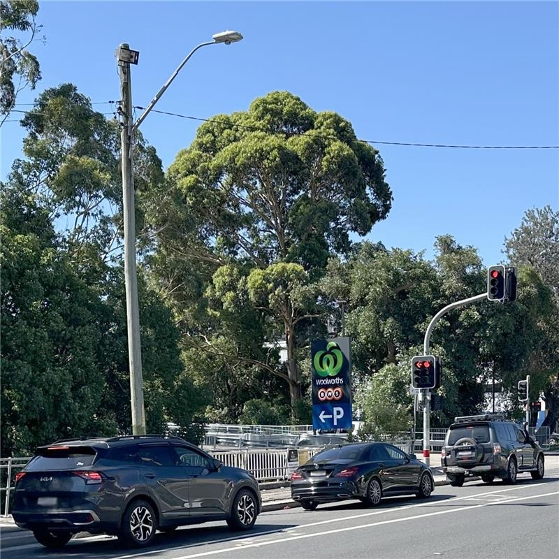 Cars wait at a red traffic light on a tree-lined street. A Woolworths parking sign is visible, and the sky is clear and blue, evoking a calm atmosphere.