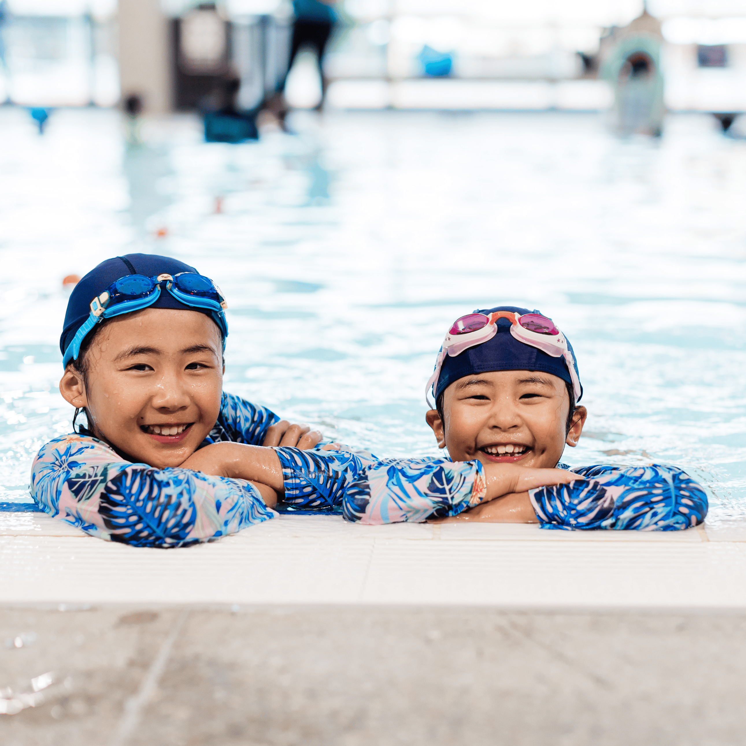 Two young children in swimwear and goggles smile joyfully at the pool's edge. The bright indoor pool environment conveys a sense of fun and play.