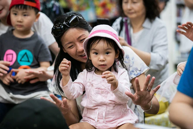 A smiling woman holds a young girl wearing a pink hat and overalls in a playful setting. Nearby children and adults look on with joyful expressions.