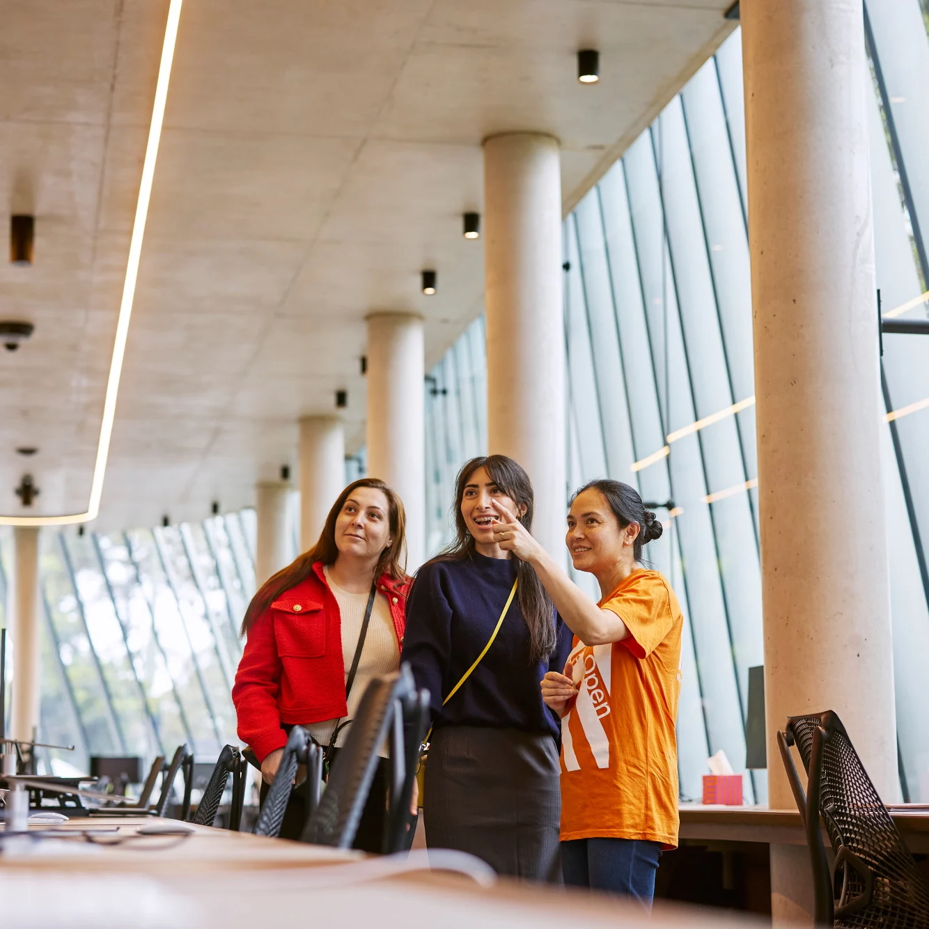 Three people standing next to desks with black chair, person in orange tshirt is pointing.