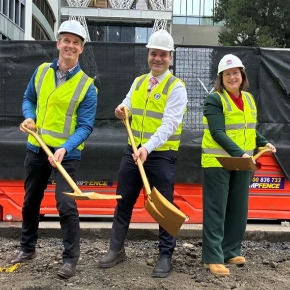 Two men and a woman in high-vis yellow vests and white hard hats hold golden shovels over dirt,