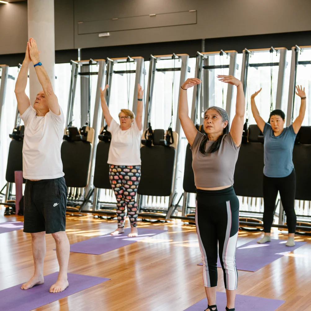 Elderly people in athletic wear stretch arms above their heads while standing on purple yoga mats.