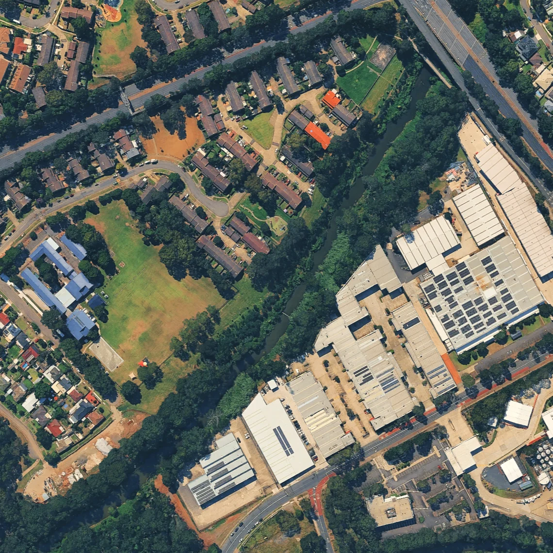 Aerial view of a landscape with residential areas on the left featuring houses and green lawns, contrasted by industrial buildings on the right, separated by a river and tree line.
