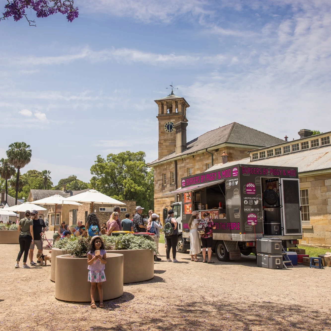 People gather around a food truck in a sunny courtyard with an old stone building and clock tower. The atmosphere is lively and relaxed.