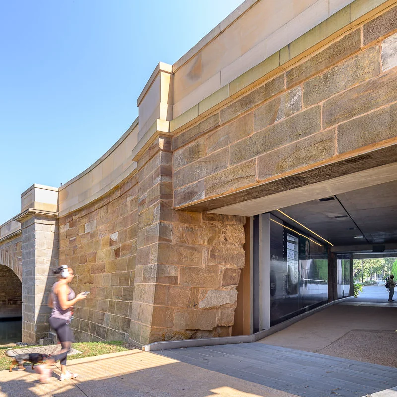 A runner holding a mobile phone runs towards a stone archway tunnel in daylight. The tunnel features modern lighting and leads to a path with distant greenery.