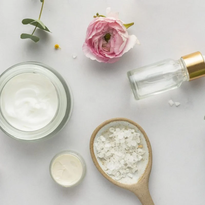 A flat lay of skincare items on a white surface. Includes an open cream jar, glass dropper, pink flower, salt in a spoon, and eucalyptus leaves. Serene vibe.