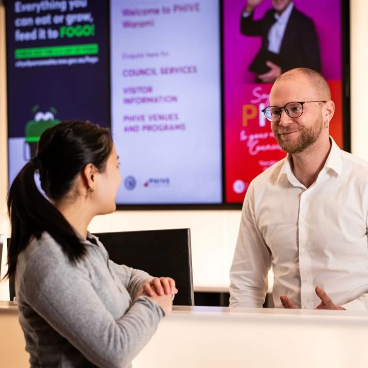 A man and woman at a reception desk engage in friendly conversation. The man, in glasses and a white shirt, smiles warmly. Digital screens are in the background.