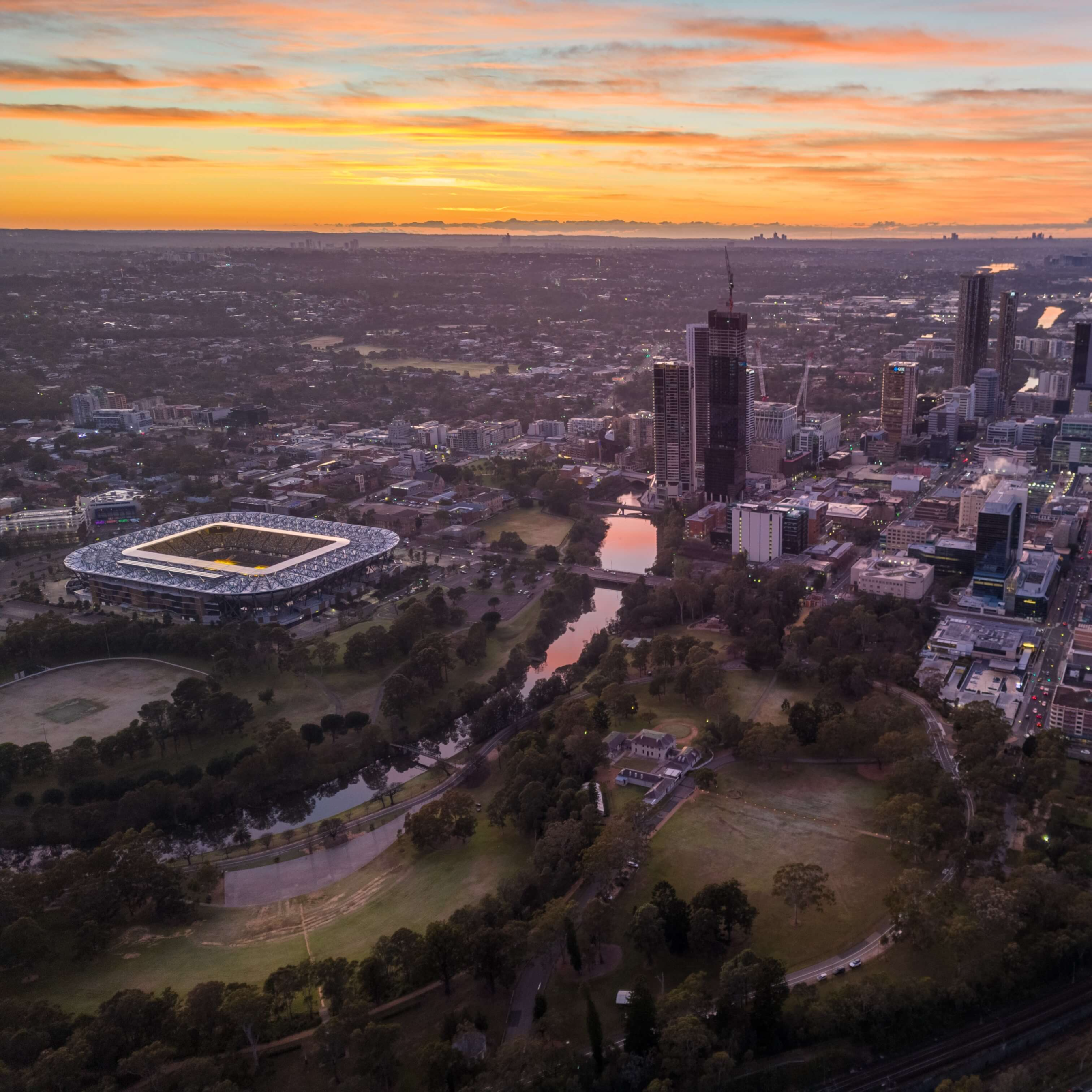 Aerial view of a city at sunset, with tall buildings and a stadium on the left. The sky is painted in soft orange and pink hues, creating a calm ambiance.