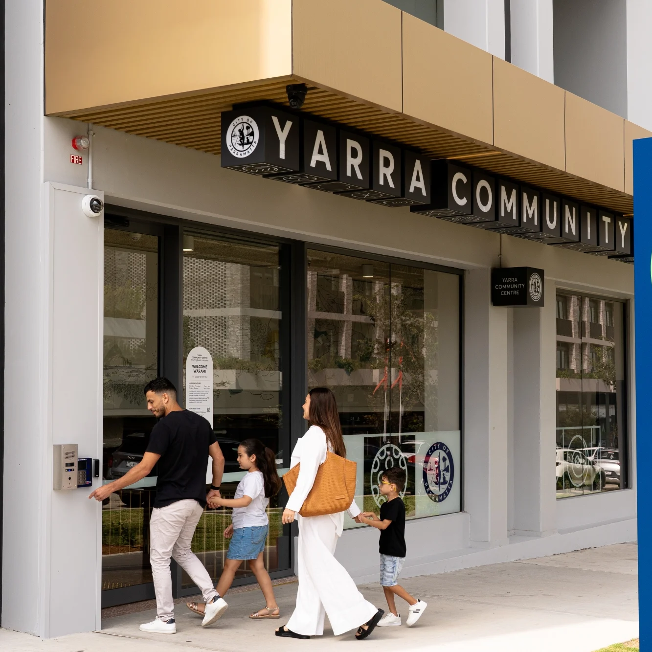 Family entering Yarra Community Centre with a modern facade. A blue sign displays the centre's name. Warm, inviting atmosphere.