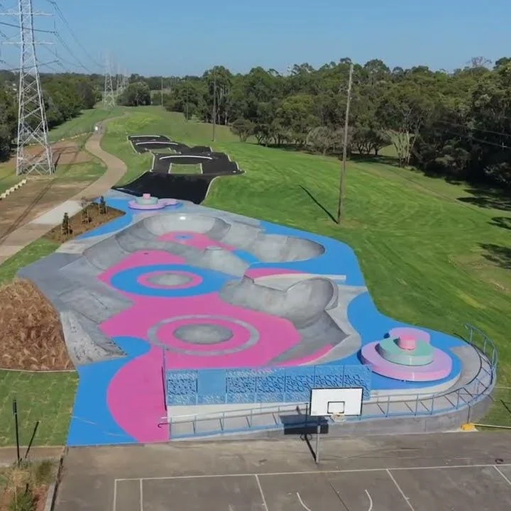 Aerial view of a colorful skatepark featuring vibrant pink and blue designs with smooth curves, set in a lush green park under a clear sky.