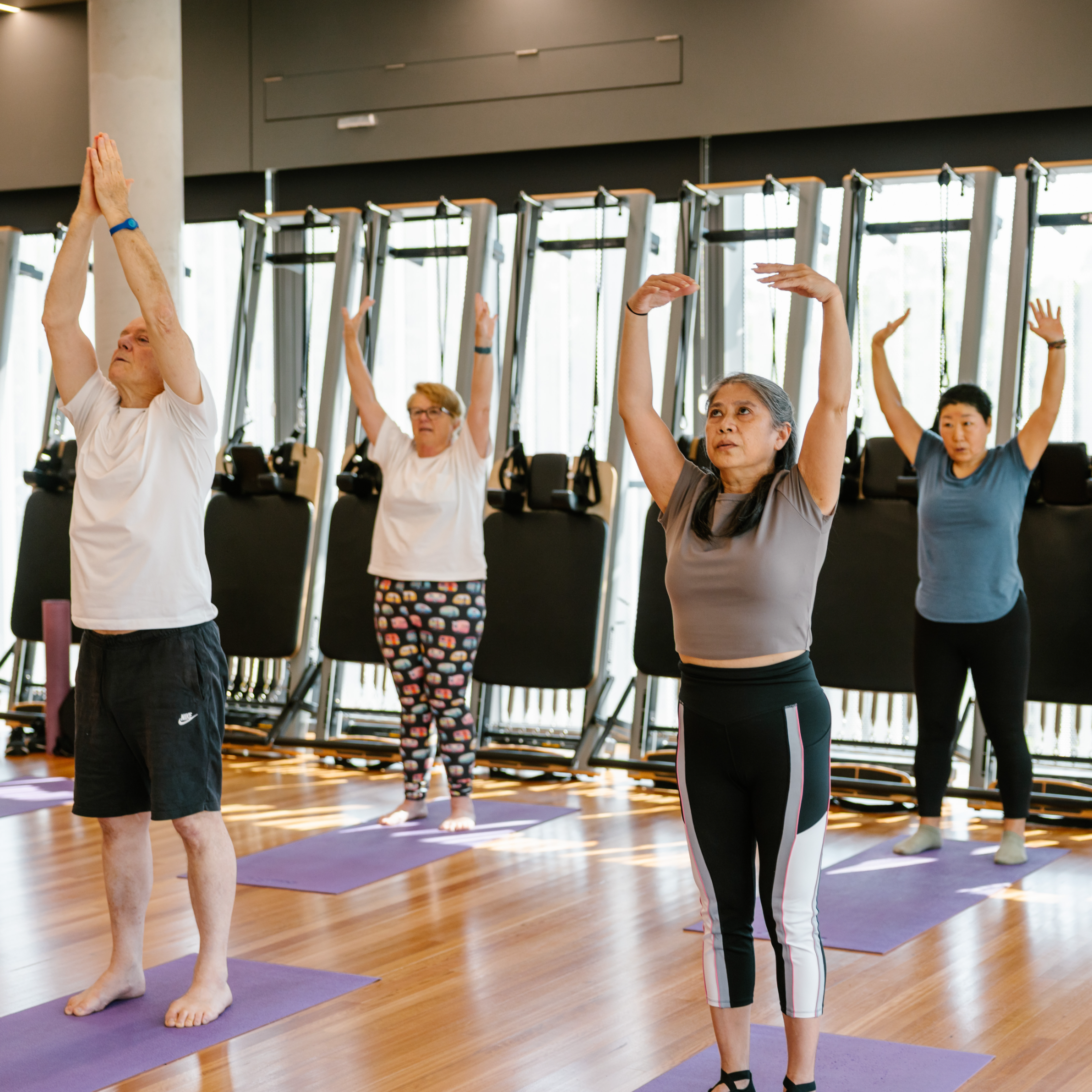 Four adults participate in a yoga class, standing on mats with arms raised, inside a gym with wooden floors and exercise equipment in the background.