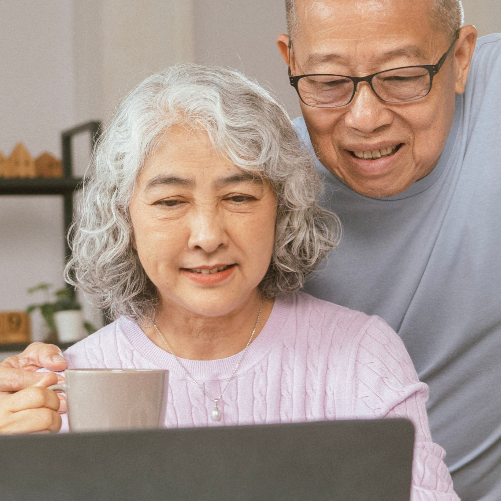 An older couple is joyfully looking at a laptop together. The woman holds a mug, suggesting a cozy atmosphere. They both wear casual clothes and glasses.