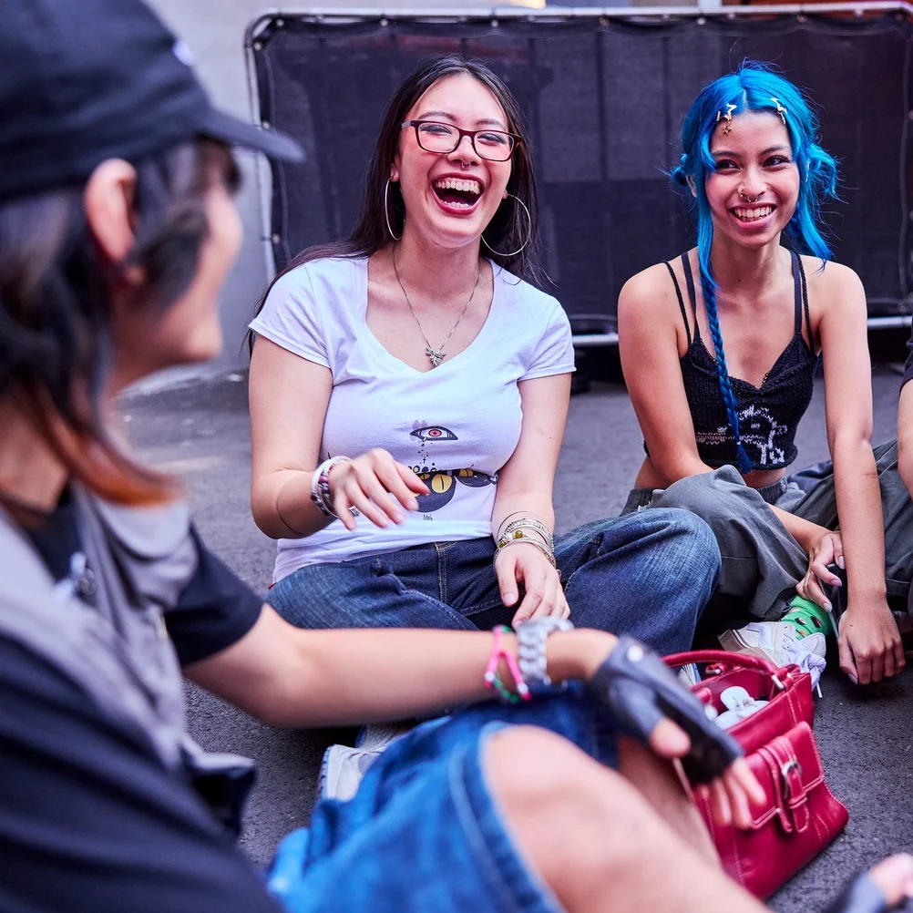 A group of four young adults, two women and two men, sit on the pavement, laughing and talking. The mood is cheerful and relaxed, with colorful attire.