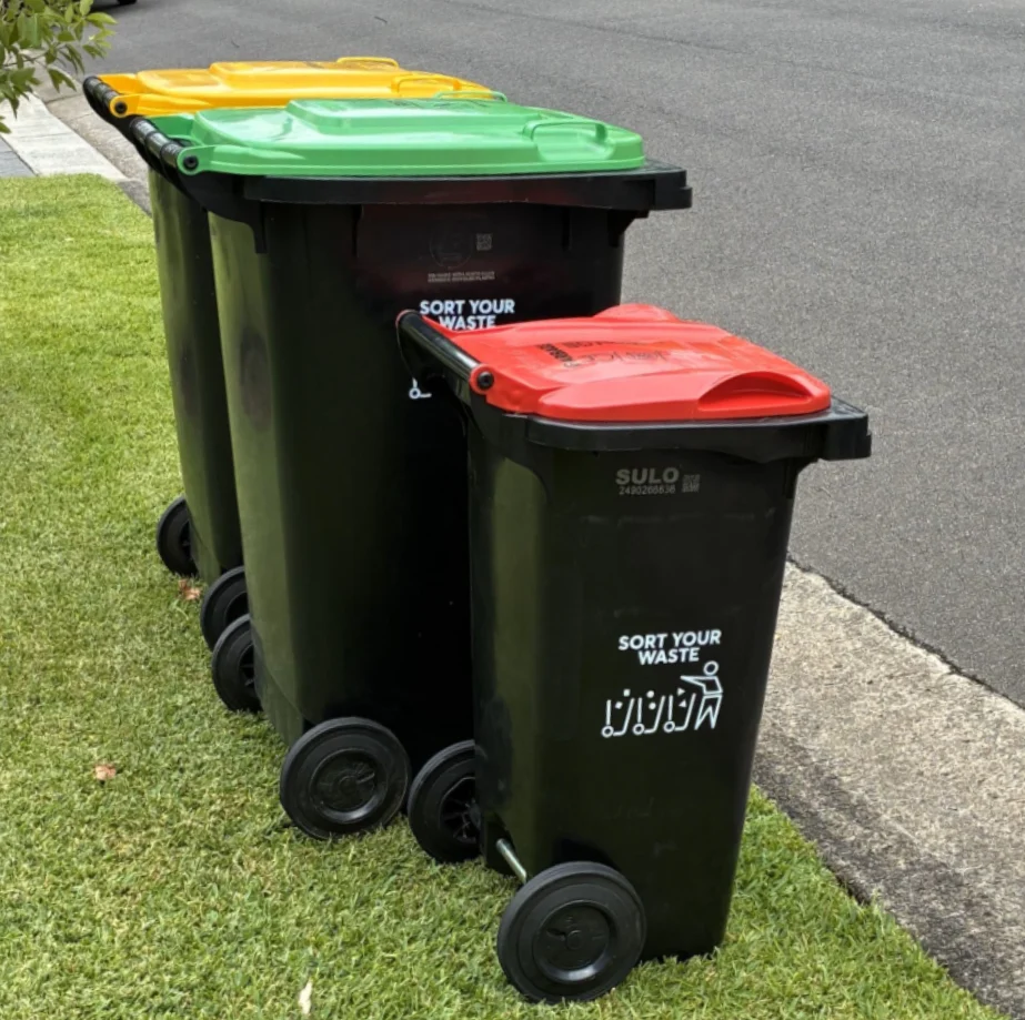 Three bins on the curb, one with a yellow lid, one with a green lid and one with a red lid.