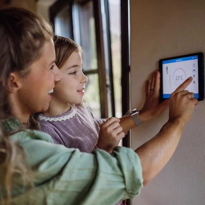 A woman and a young girl smile while adjusting a smart thermostat mounted on a wall. The display shows 23°C, setting a warm, cozy atmosphere.