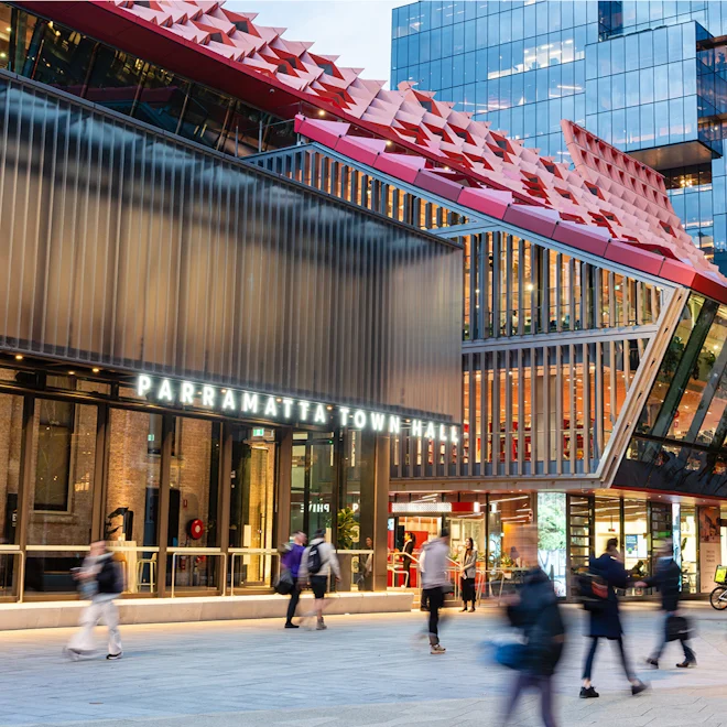 Modern city scene at dusk outside Parramatta Town Hall, featuring a sleek glass facade, vibrant red design elements, and blurred pedestrians conveying motion.