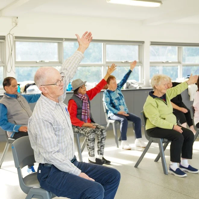 Elderly people sitting in grey chairs holding arms in the air