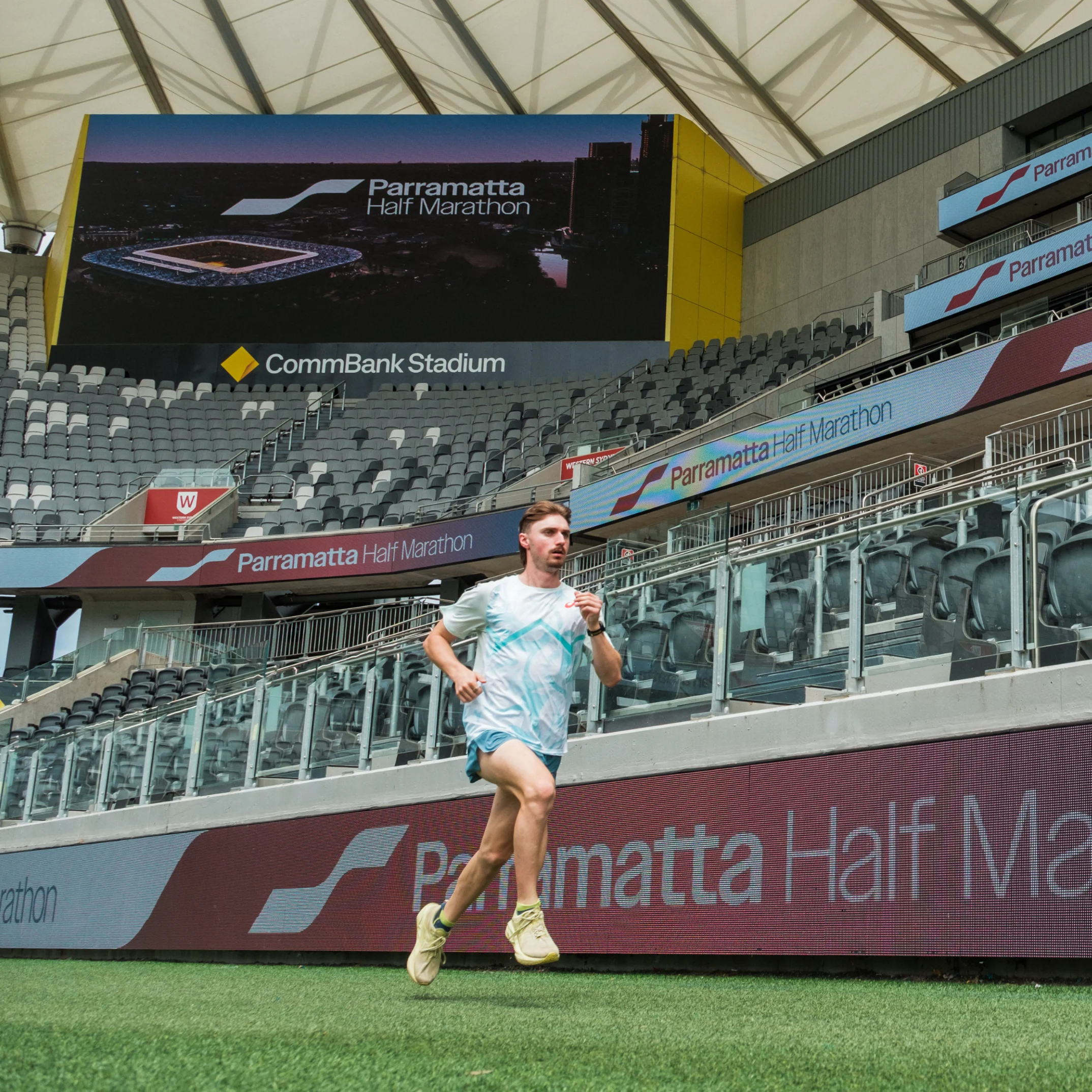 A runner in light athletic gear jogs on a stadium track. Empty bleachers and Parramatta Half Marathon signs create a focused, determined mood.