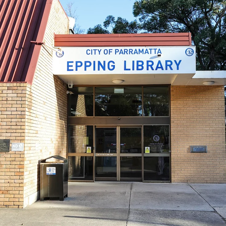 Front view of Epping Library entrance in Parramatta. Brick building with a large sign, glass doors, outdoor greenery, and a peaceful ambiance.