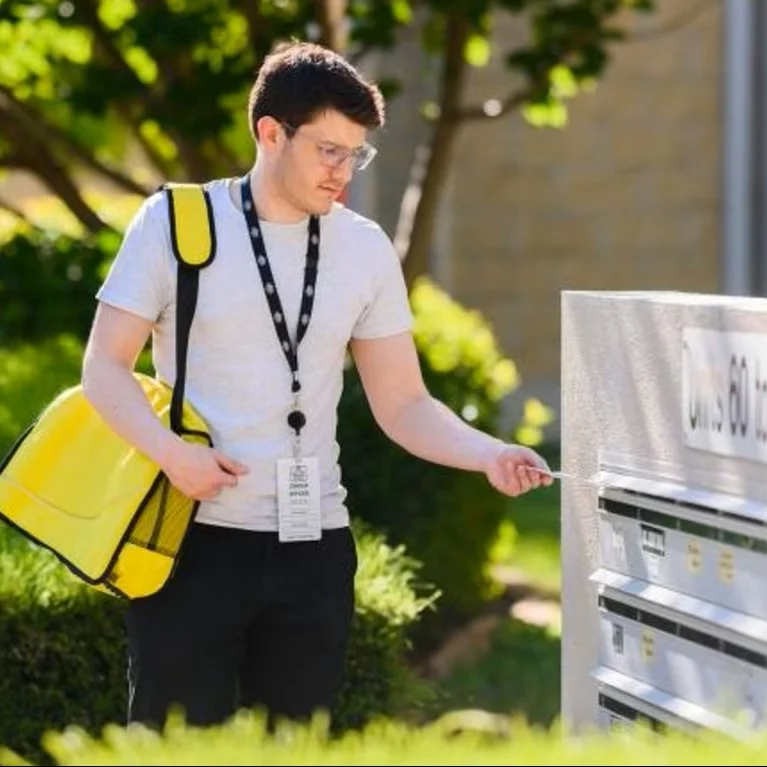 A delivery person wearing a white shirt and glasses uses a key to open a mailbox. They carry a bright yellow bag. The setting is a sunny day with greenery.