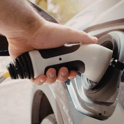 A hand plugs an electric vehicle charger into a car, emphasizing eco-friendly technology. Sunlight filters through trees, creating a warm, sustainable mood.