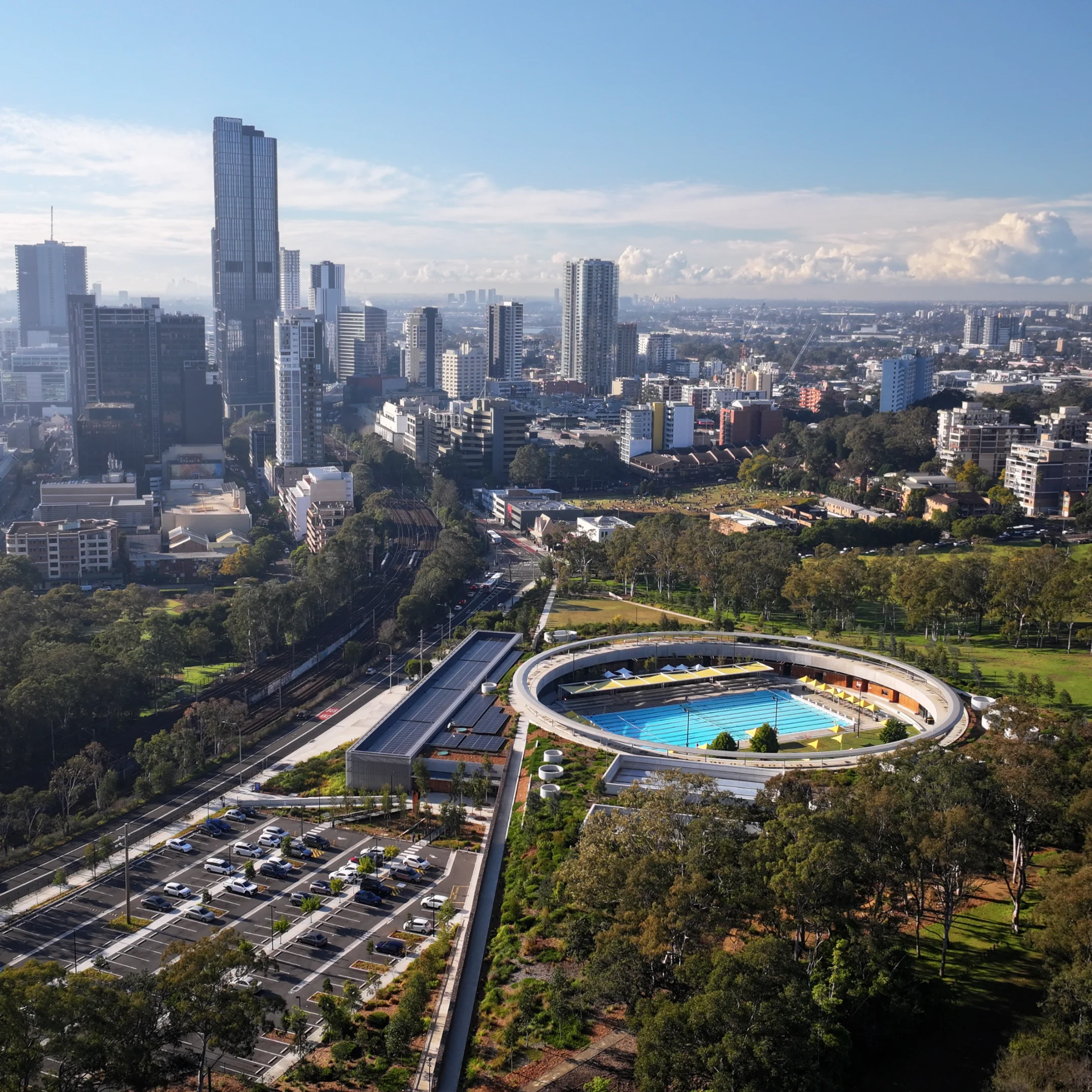 Aerial view of a city skyline with tall buildings under a blue sky. In the foreground, a circular swimming pool is surrounded by trees and parking lot.