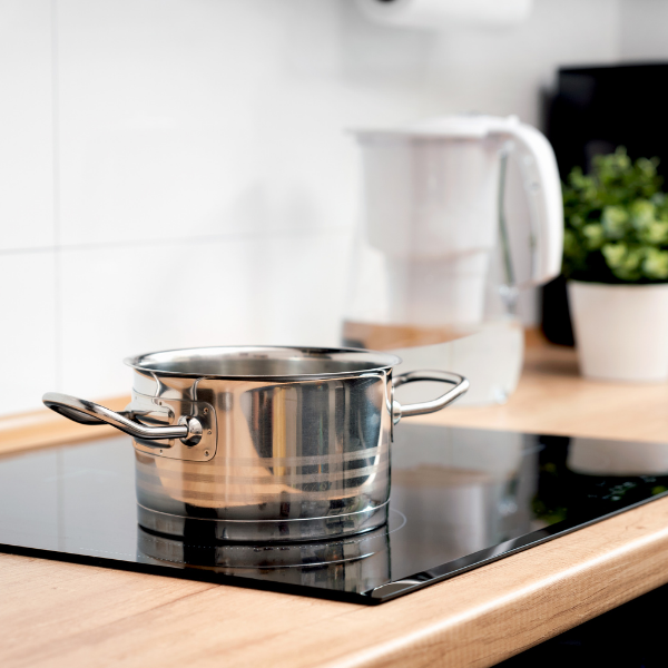 A shiny stainless steel pot sits on a modern induction stove. In the background, a white water filter pitcher and a small potted plant add a fresh touch.