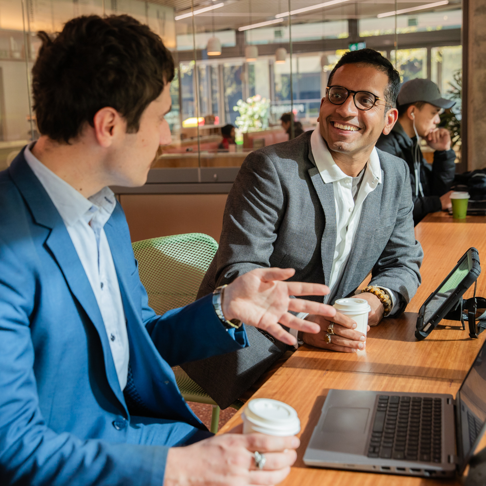 Two men in business attire converse at a bright, modern café, each holding coffee cups. They sit at a wooden table with laptops, smiling warmly.