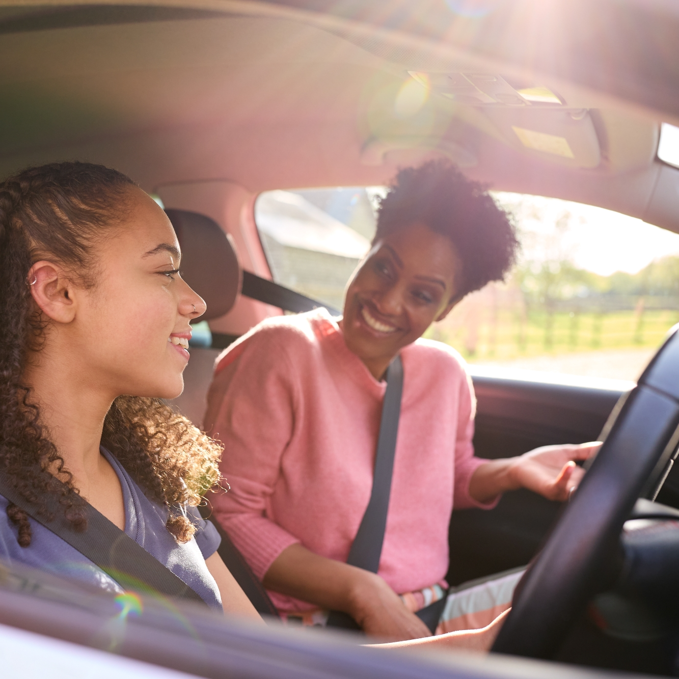 A young woman driving a car, smiling, with an older woman in the passenger seat, also smiling. Sunlight streams through the window, creating a warm, positive atmosphere.