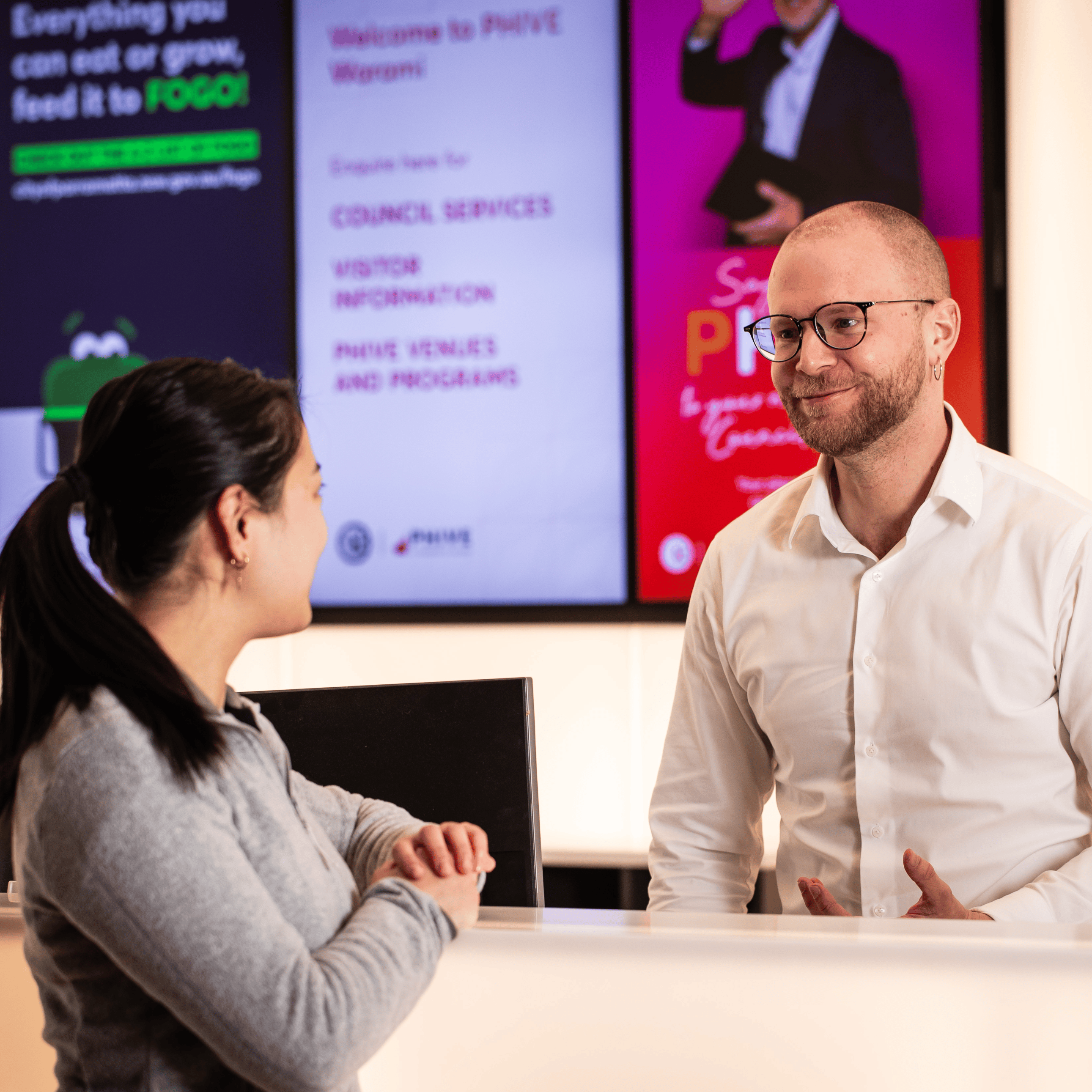 A receptionist in a white shirt smiling and talking to a woman at a counter. Bright digital signs are visible in the background, creating a welcoming atmosphere.