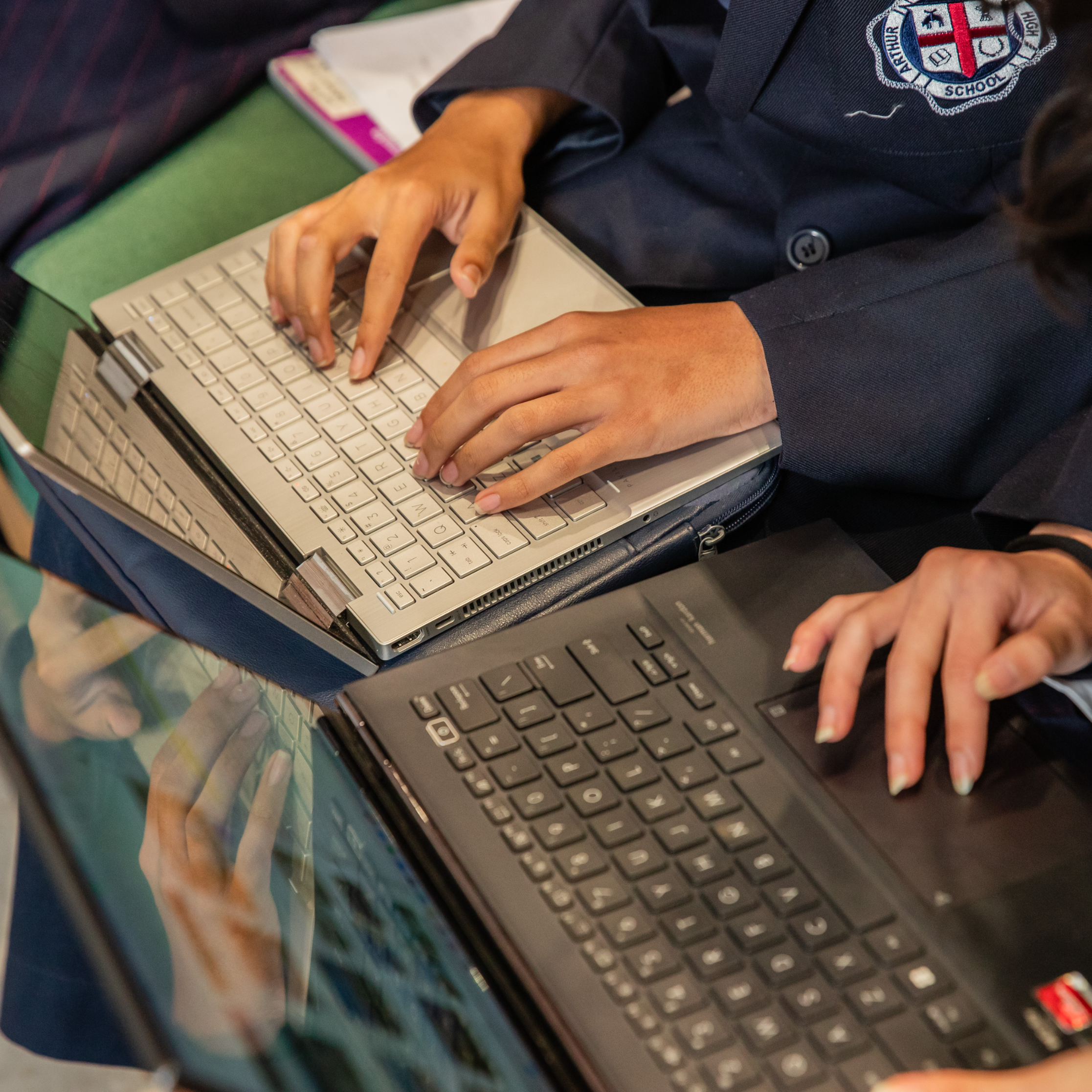 Students in uniforms type on laptops, displaying focused collaboration. Their badges and formal attire suggest a school setting, creating a studious atmosphere.