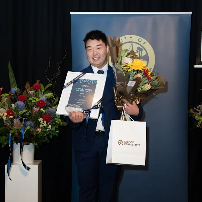 Man in a suit holding a "Young Citizen of the Year" award, a bouquet, and a City of Parramatta bag. He stands smiling against a dark backdrop with floral arrangements.