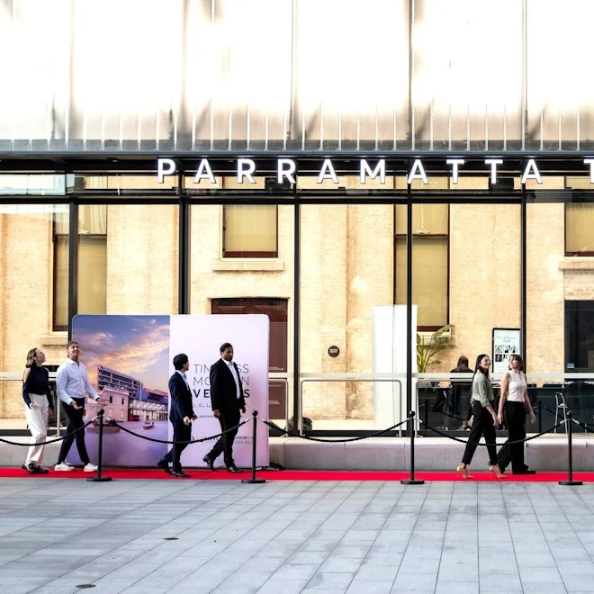 People walk on a red carpet outside a modern building with "Parramatta" signage. The atmosphere is formal and contemporary.