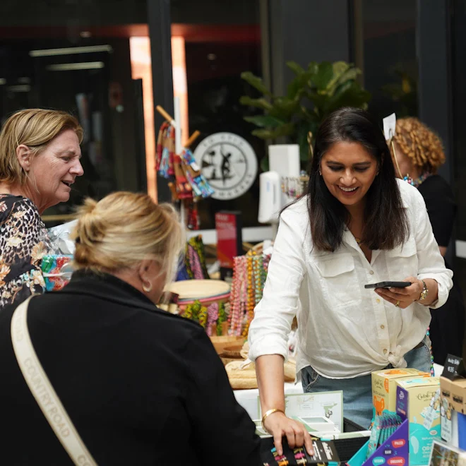 A woman in a white blouse joyfully interacts with customers at a vibrant market stall with colorful necklaces and boxes. The atmosphere is lively and friendly.