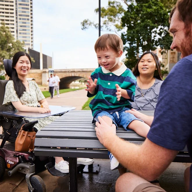 A joyful child in a striped shirt sits on a park table, surrounded by smiling friends. One person is in a wheelchair. The scene is lively and inclusive.