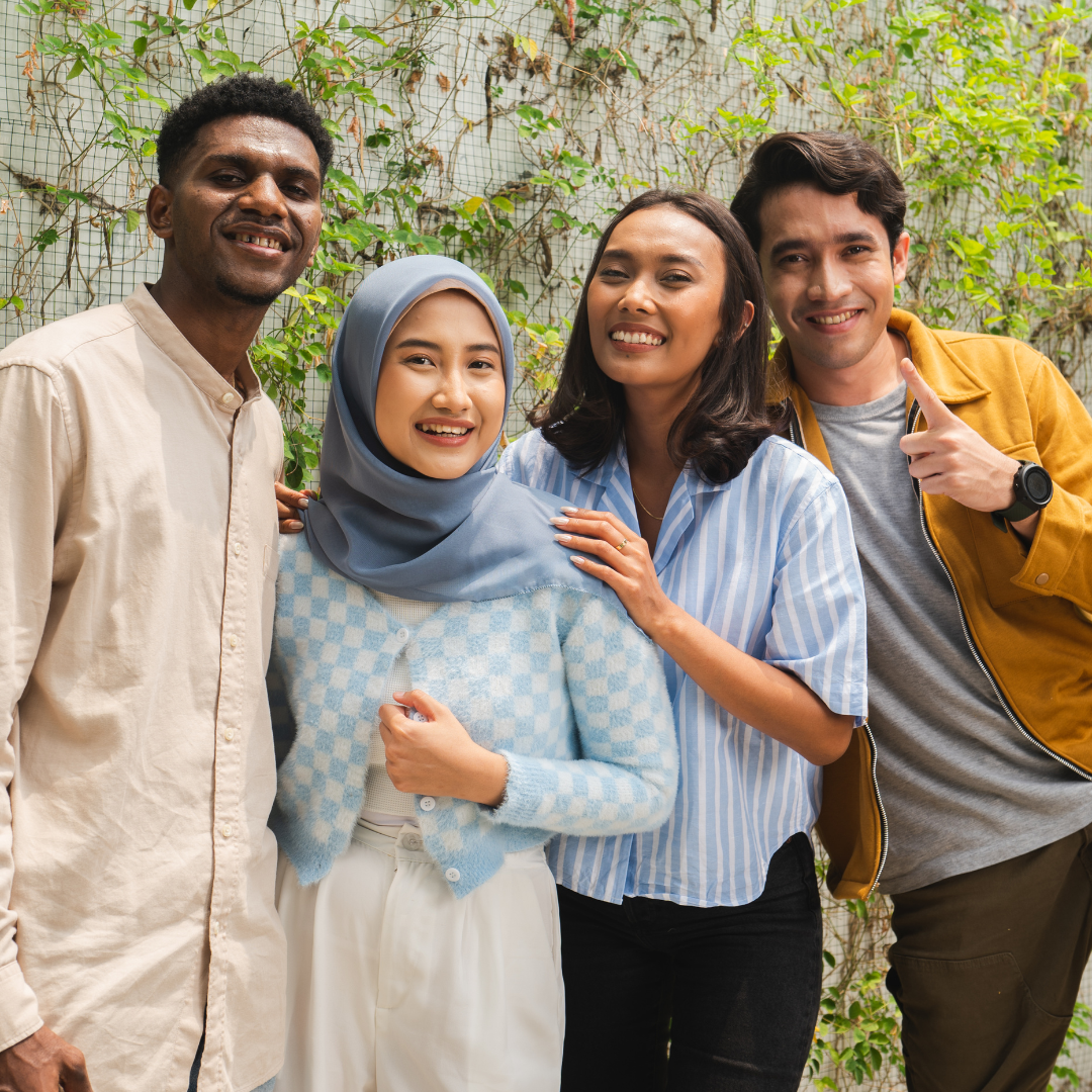 Four friends smiling in a garden. One wears a hijab, and another points playfully. They exude warmth and happiness against a lush green backdrop.