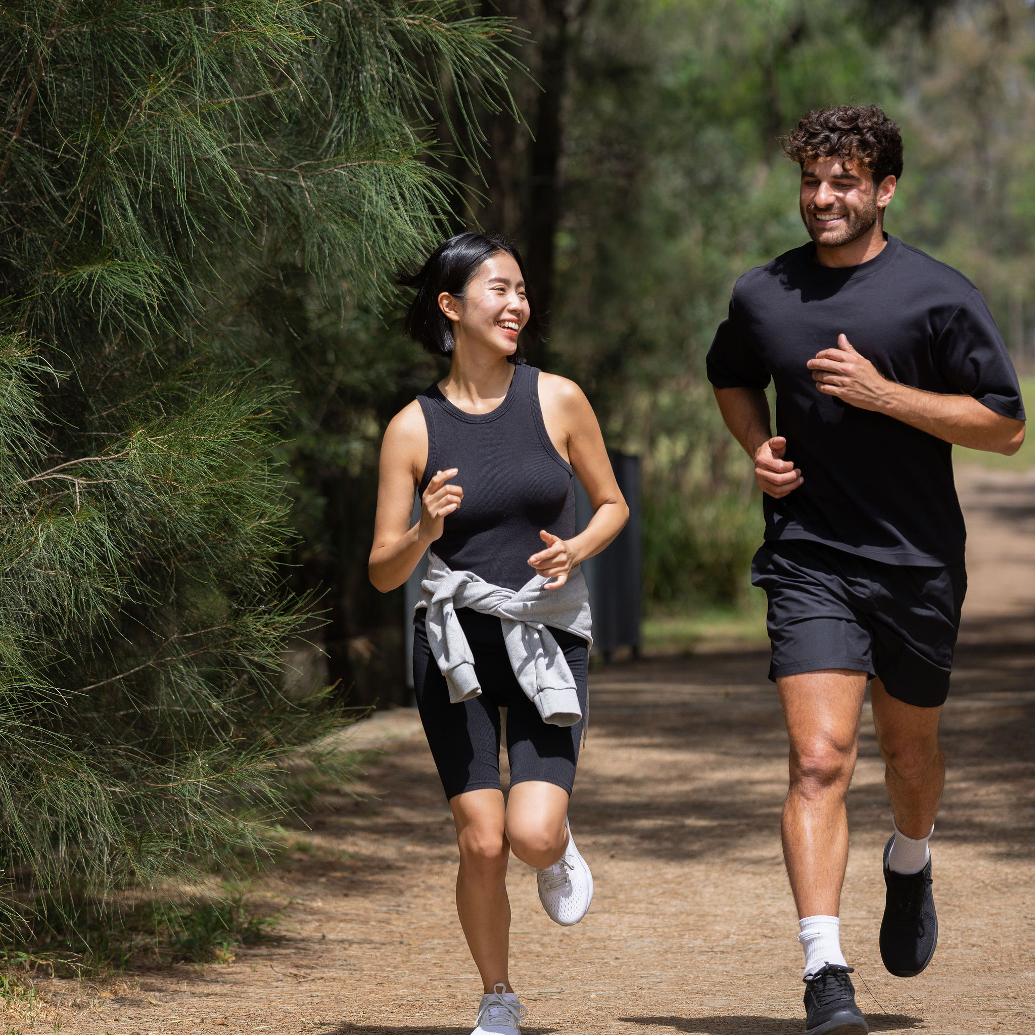 A man and woman jogging on a wooded path, smiling and talking. They wear athletic gear, conveying a sense of joy and camaraderie in a natural setting.