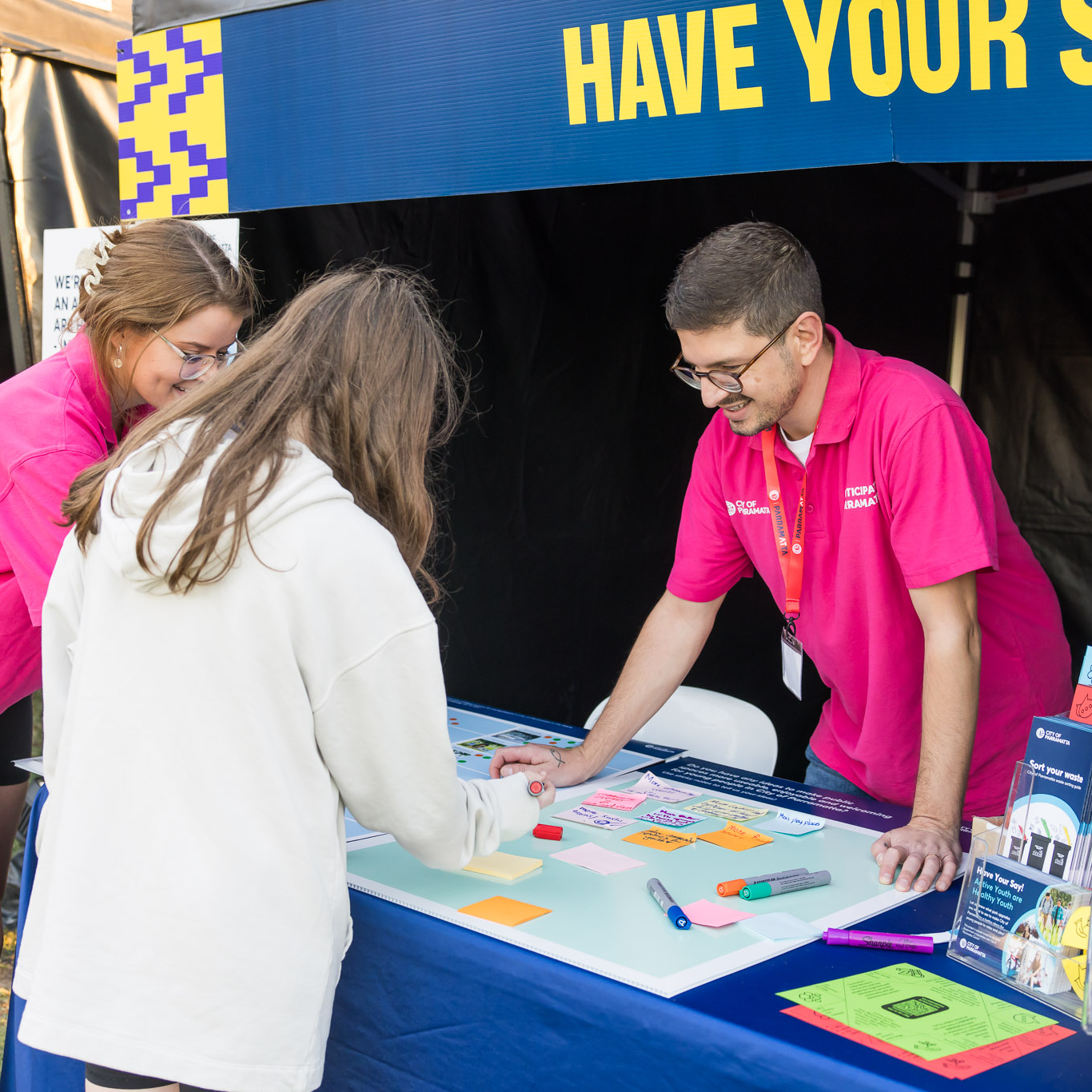 A community engagement booth with a "Have Your Say" banner. Two volunteers in pink shirts assist a woman writing on a board with colorful sticky notes. The atmosphere is welcoming and inclusive.