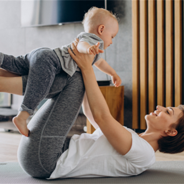 A woman lies on a yoga mat indoors, lifting a smiling baby in the air with her legs. The scene is bright and playful, conveying a sense of joy and bonding.