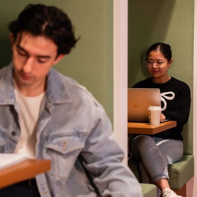 A woman in glasses works on a laptop in a cozy green cubicle, holding a coffee cup. In the foreground, a man in a denim jacket reads attentively.