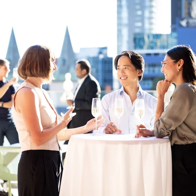 Three people stand outside around a table with drinks, engaged in a lively conversation and smiling. City buildings are visible in the bright background.