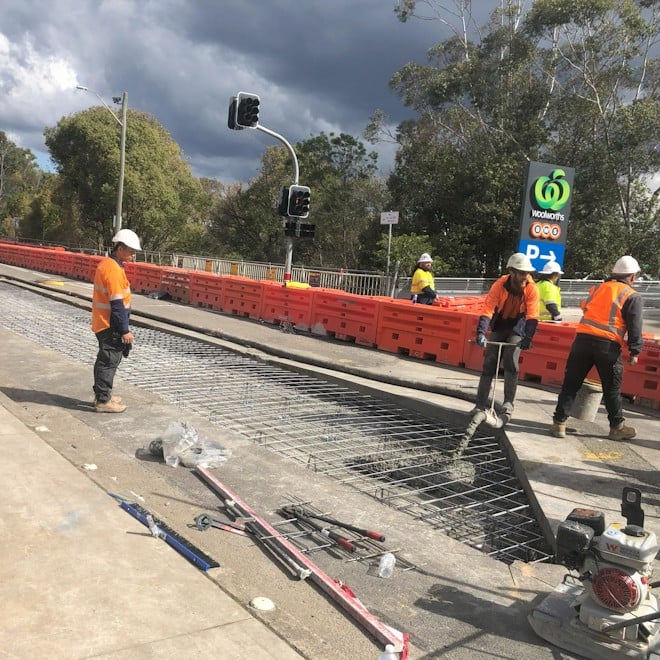 Construction workers in orange safety gear lay concrete on a road site by Woolworths. Orange barriers and overcast skies are visible in the background.