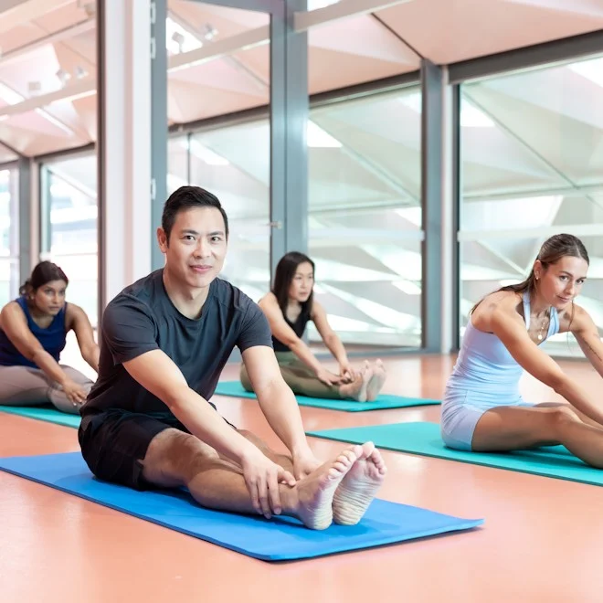 Four individuals on yoga mats perform seated forward bends in a bright, modern studio. They exhibit focus and calm, with natural light streaming in.