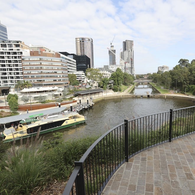 Riverside scene with modern buildings, greenery, and a ferry docked on the river. Overcast sky adds a calm, urban atmosphere.