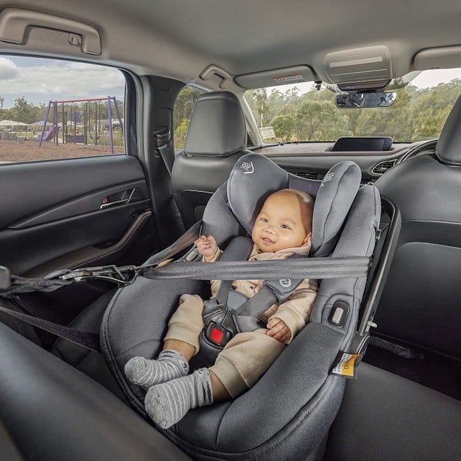 A smiling baby in a gray car seat, safely buckled, sits in the backseat of a car. A swing set is visible outside the window, indicating a park setting.