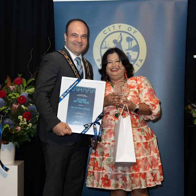 Man and woman smiling at award ceremony. The man holds a framed "Citizen of the Year" certificate, and the woman holds a trophy. Background has flowers and city emblem.