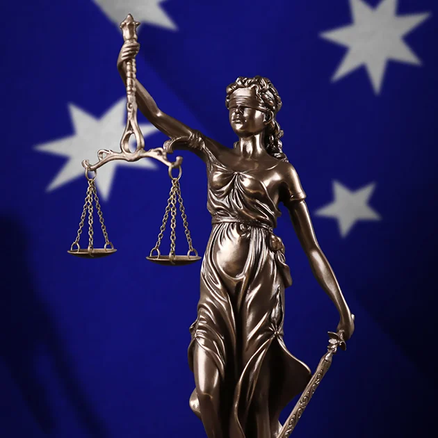 Bronze Lady Justice holds balanced scales and a sword before a blurred Australian flag, evoking fairness, law, and national identity.
