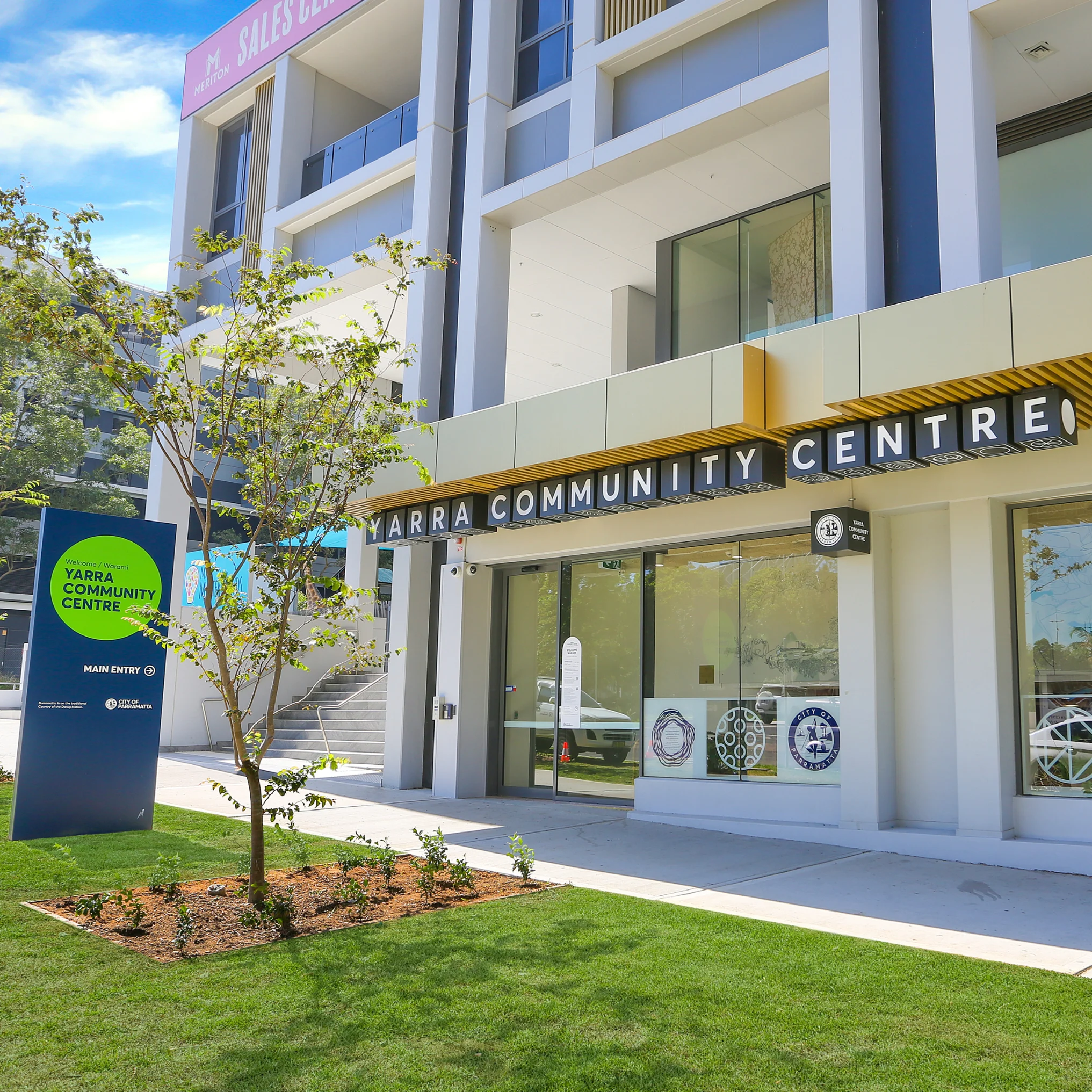 Sunlit façade of the modern community centre, with large glass windows, a yellow slatted awning, trees, and a green lawn; bright, welcoming feel.